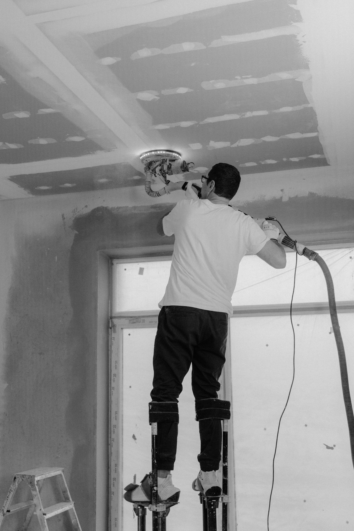 Worker on drywall stilts running a sander with LED light ring across a taped ceiling, shown in black and white