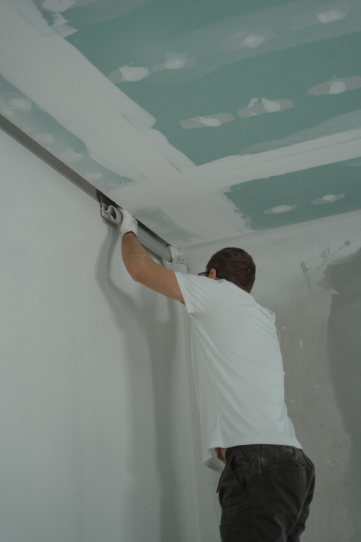 Worker pressing a wide finishing knife along the joint where a drywall ceiling meets the wall