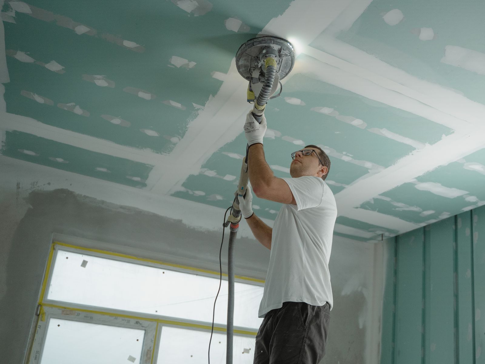 Tradesman using a vacuum-attached ceiling sander to level fresh joint compound on a drywall ceiling
